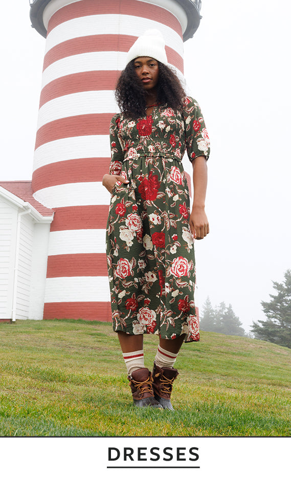 A model stands outdoors in front of a red-and-white striped lighthouse wearing a knee-length floral dress with short sleeves, a white beanie, striped socks, and brown lace-up boots. The word 'DRESSES' appears in a text block at the bottom.