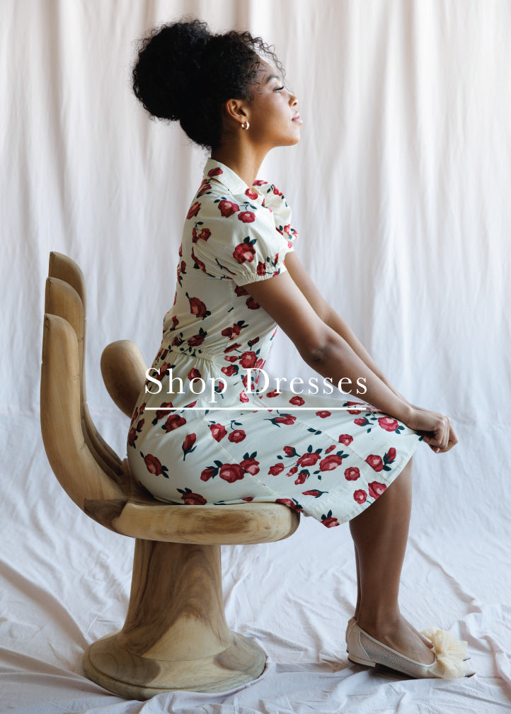 Woman sitting on a wooden chair wearing a floral dress with text 'Shop Dresses' overlayed.