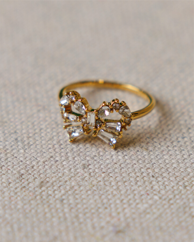 Gold bow-shaped ring with cubic zirconia gemstones on a textured beige surface