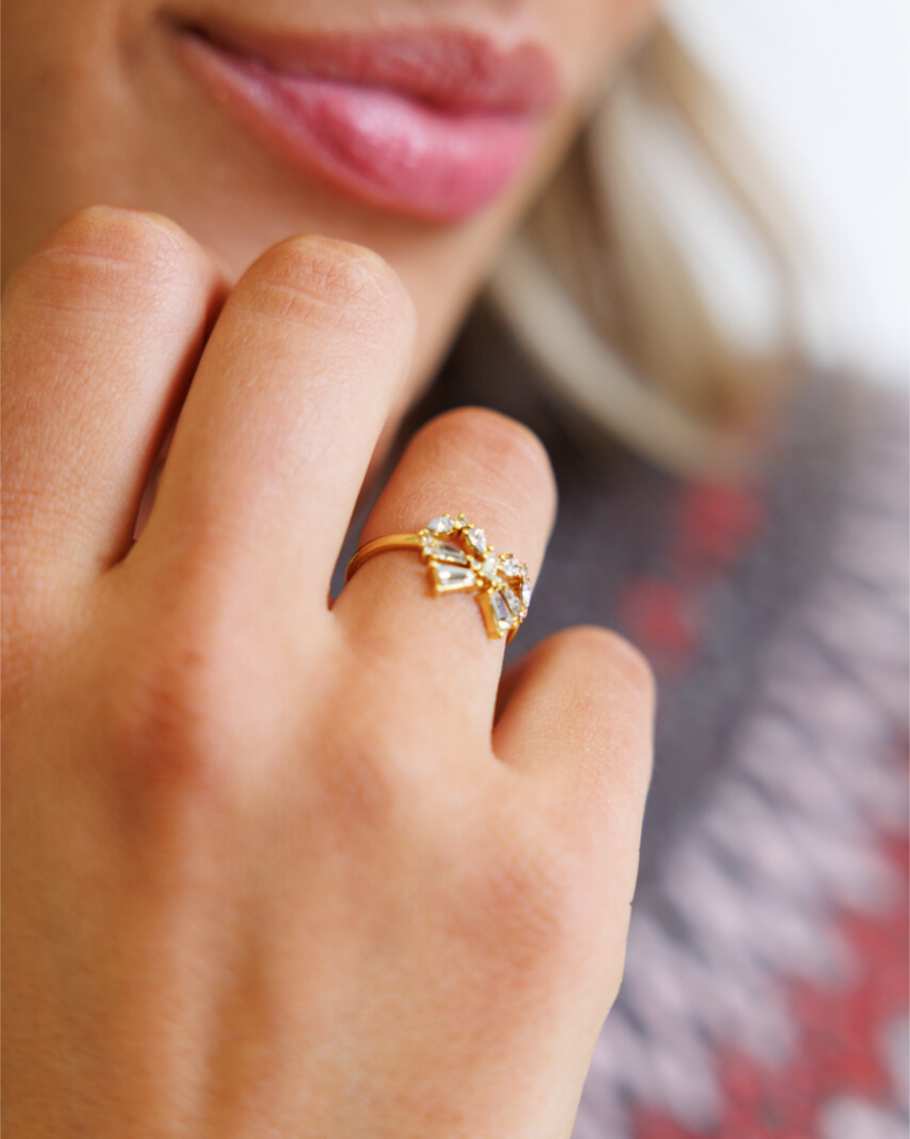 Close-up of a hand wearing a gold bow-shaped ring with diamond gemstones, against a blurred background.