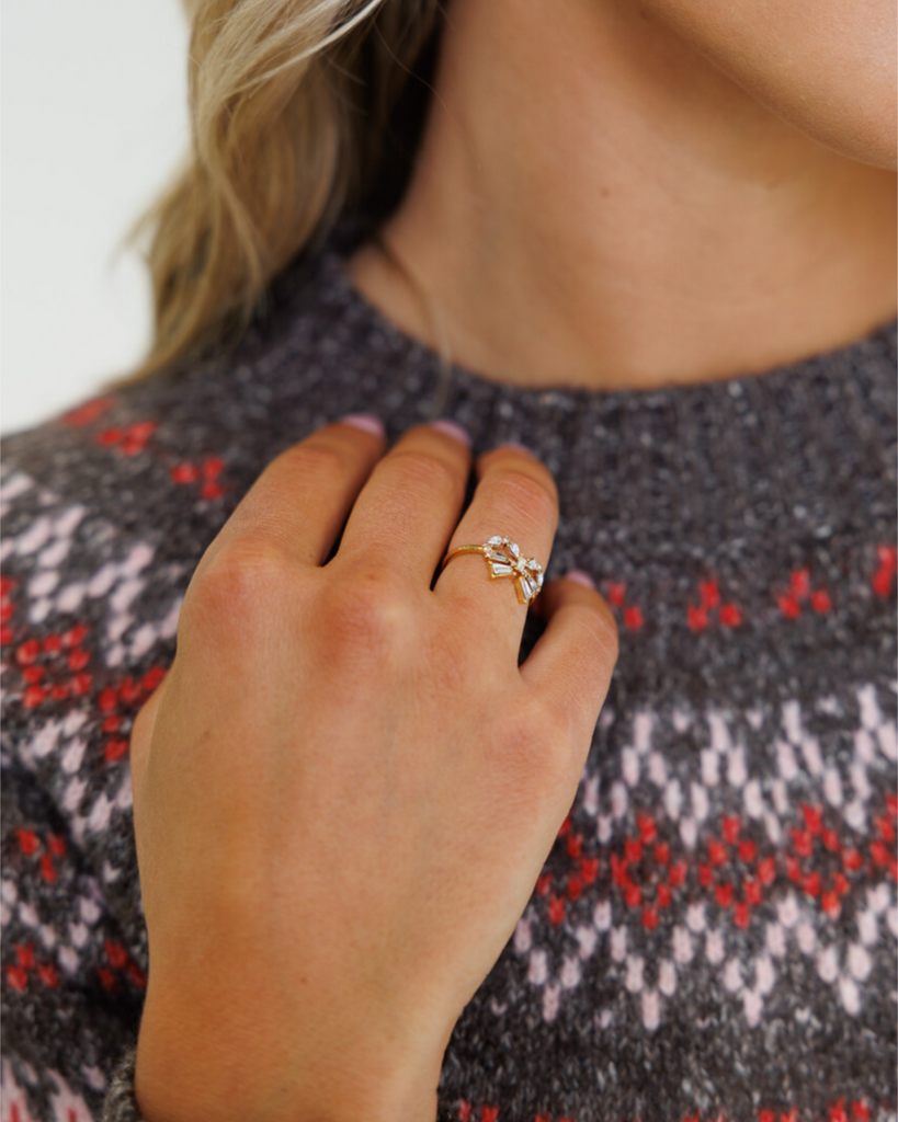 Close-up of a hand wearing a gold bow-shaped ring with diamond gemstones, against a patterned sweater background.