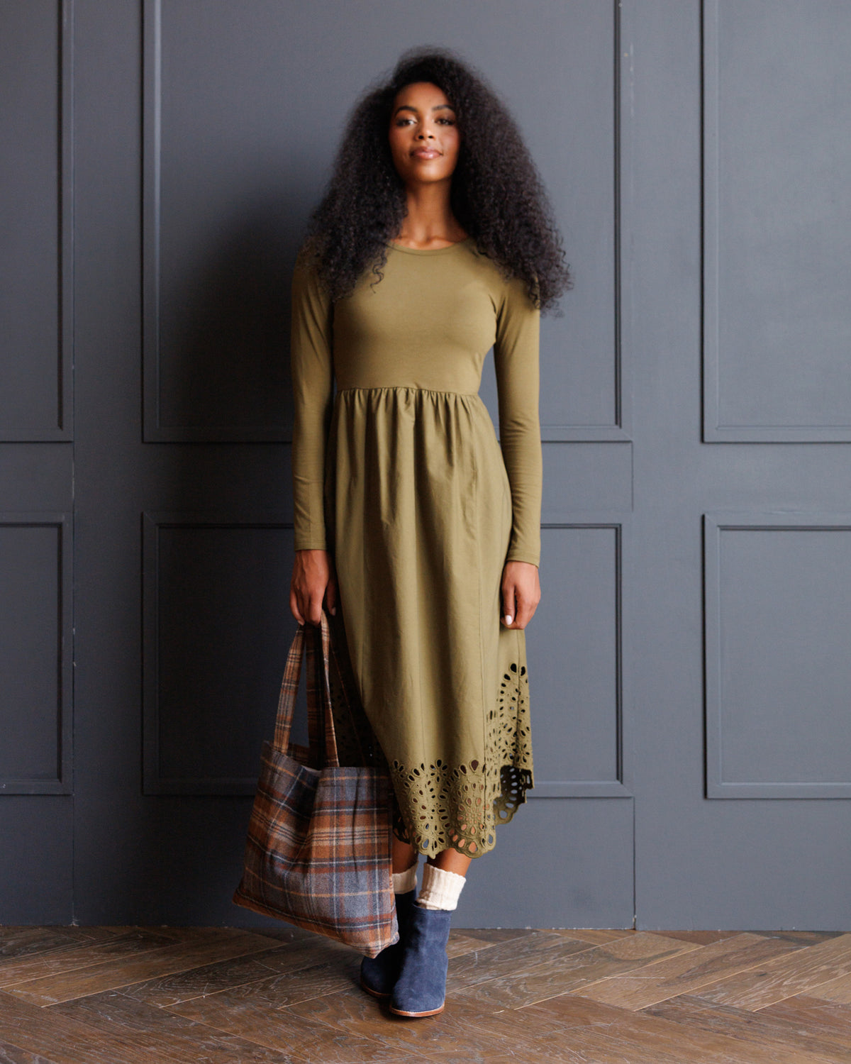 A woman stands against a dark gray paneled wall wearing a long-sleeve olive green midi dress with a gathered waist and a lace-up hem. She is holding a plaid shopping bag and wearing blue boots.