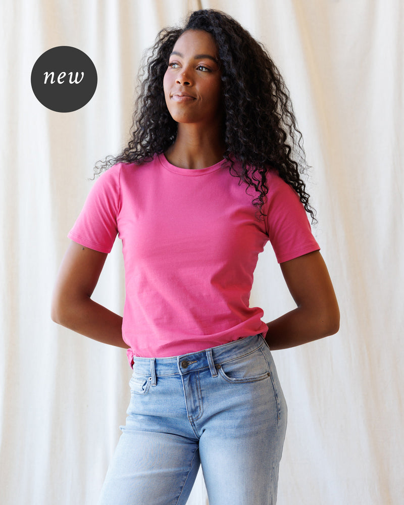 A woman wearing a pink short-sleeve crewneck t-shirt and light blue jeans, standing against a fabric backdrop.