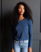 A woman with long, curly black hair wearing a fitted, long-sleeve blue crewneck top and light wash blue jeans, standing against a dark gray paneled wall.