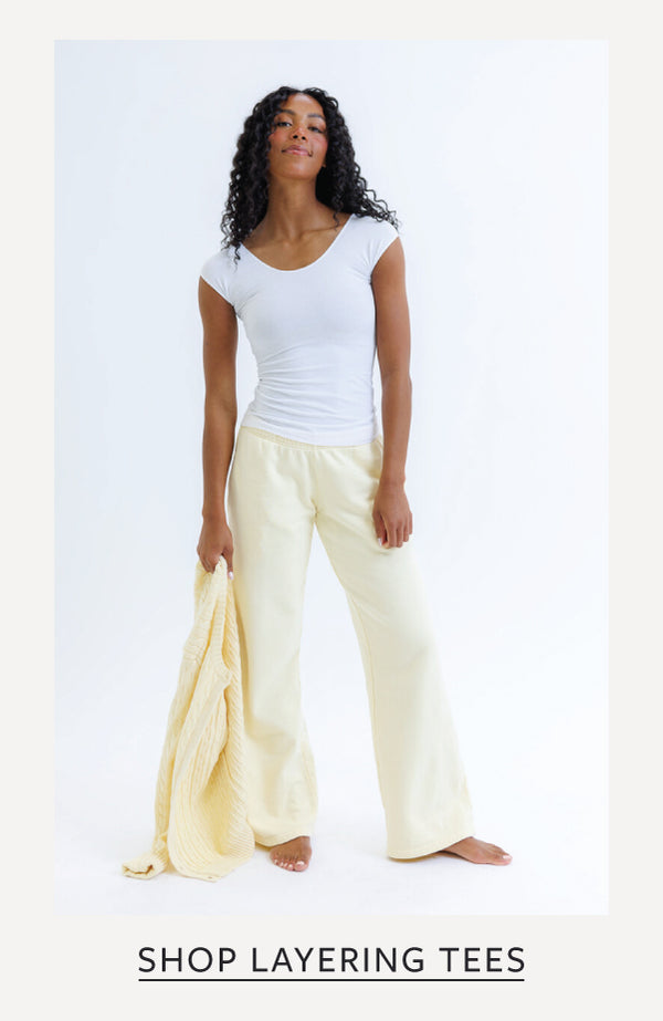 A woman stands barefoot against a plain white studio background wearing a fitted white short-sleeve top and pale cream wide-leg pants, while holding a matching cream fabric drape. Text at the bottom reads 'SHOP LAYERING TEES'.