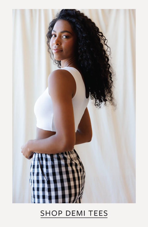 A woman with long curly black hair is shown from the back wearing a white sleeveless crop top and black-and-white gingham pants, standing against a cream curtain backdrop. Text at the bottom reads 'SHOP DEMI TEES'.