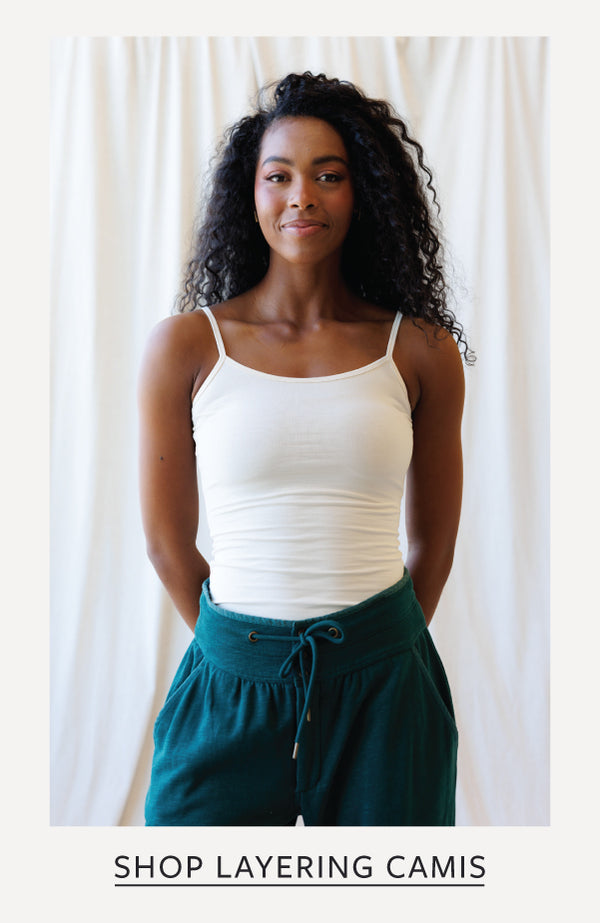 A woman with curly dark hair wearing a white camisole tank top and dark teal drawstring pants, standing against a light cream curtain backdrop. Text at the bottom reads 'SHOP LAYERING CAMIS'.