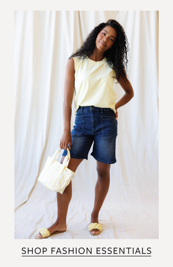 A woman modeling a pale yellow sleeveless top and dark blue denim shorts, holding a white tote bag, against a draped cream fabric backdrop. Text at the bottom reads 'SHOP FASHION ESSENTIALS'.