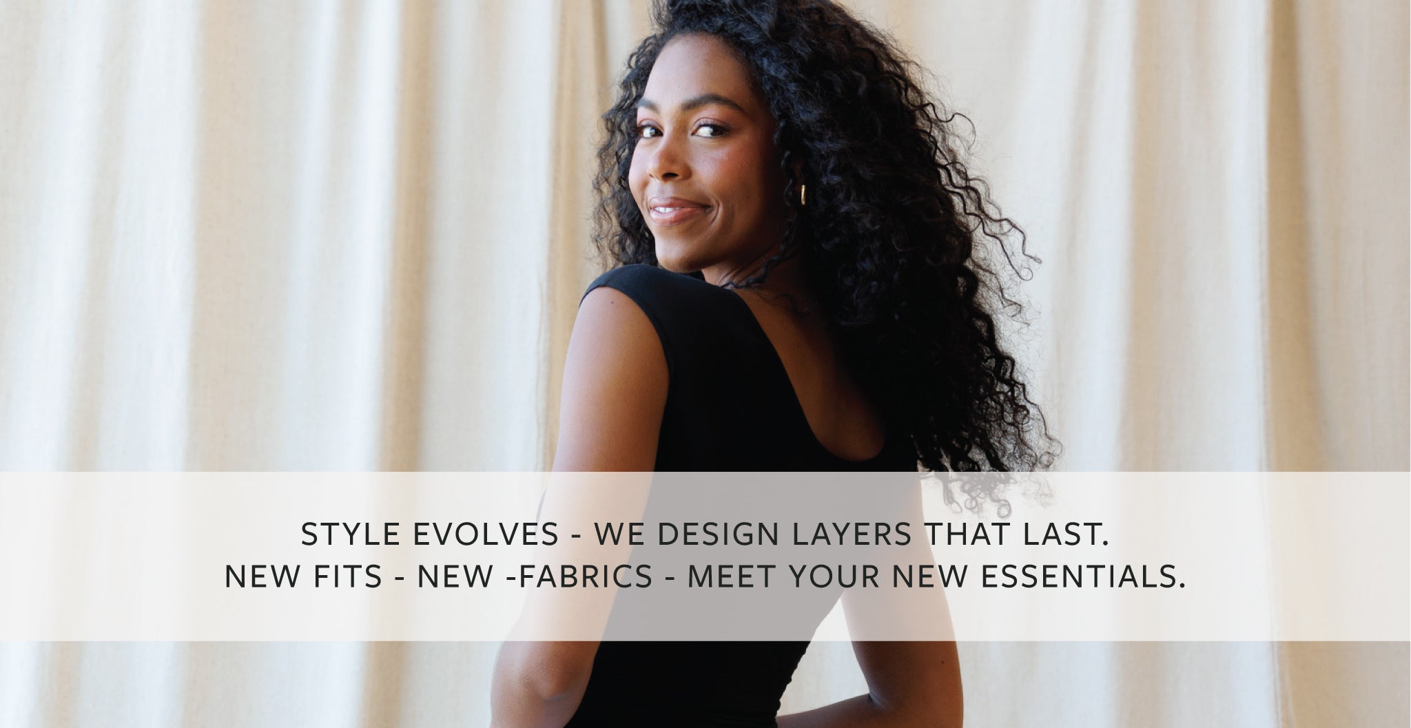 A woman with long curly dark hair is shown from the back wearing a black sleeveless top, standing in front of cream-colored curtains. A semi-transparent white banner across the bottom contains the text: 'STYLE EVOLVES - WE DESIGN LAYERS THAT LAST.