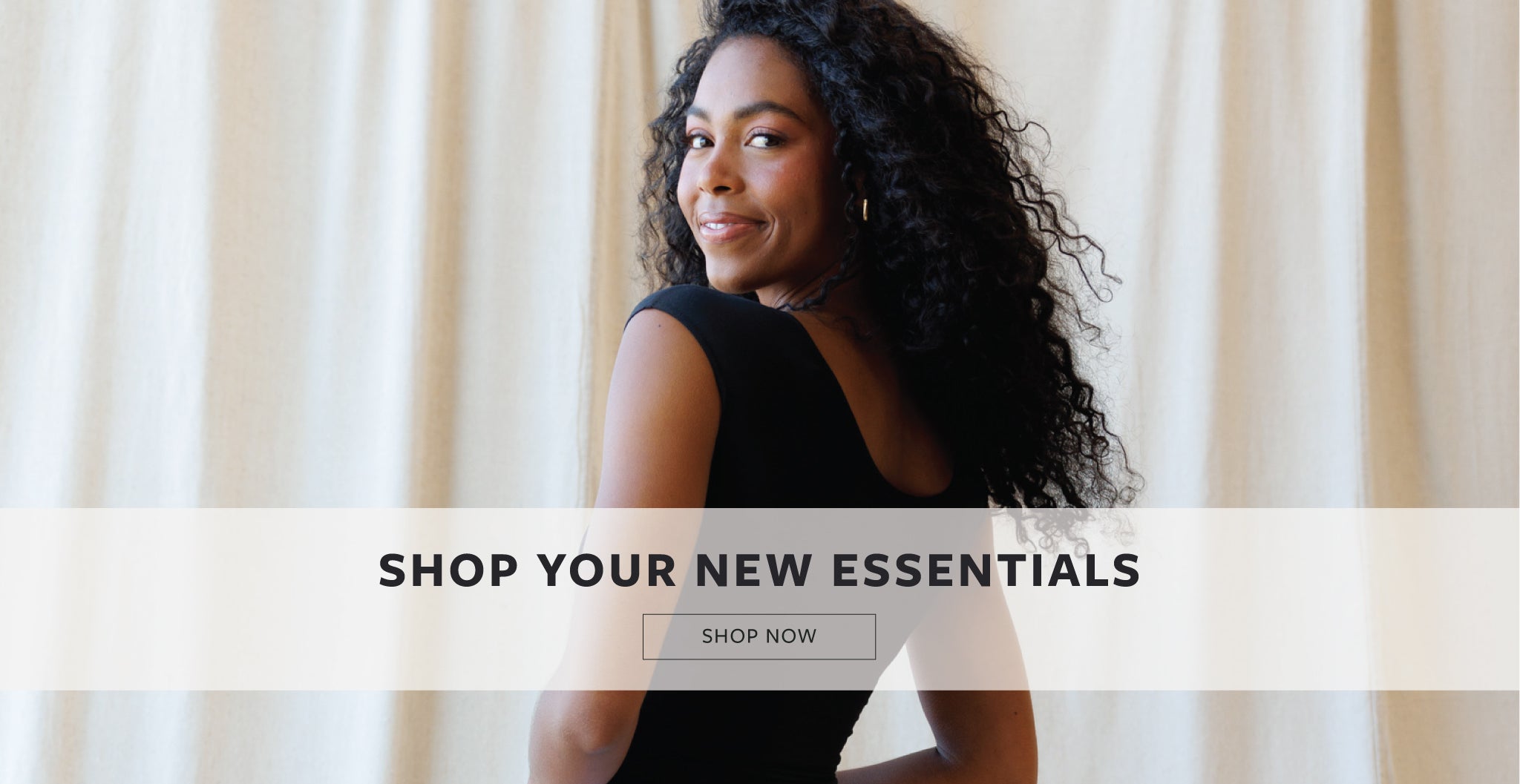 A woman with curly black hair looks back over her shoulder while wearing a black sleeveless top, standing in front of a soft beige curtain background. A white banner across the lower half reads 'SHOP YOUR NEW ESSENTIALS' with a 'SHOP NOW' button below.