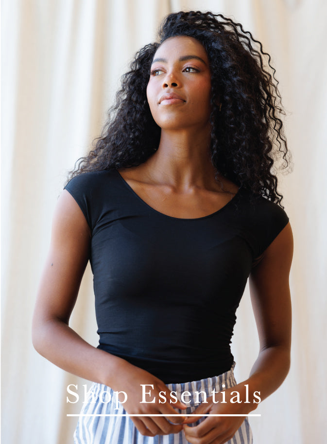 A woman with long, curly black hair wearing a fitted black scoop-neck short-sleeve top and striped pants, standing against a light beige curtain background. The text 'Shop Essentials' is overlaid at the bottom.