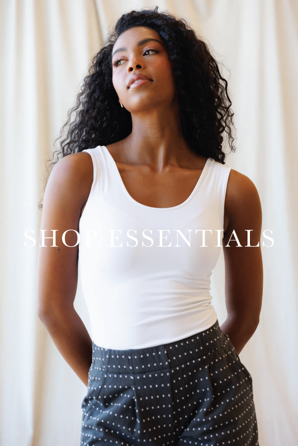 A woman with curly black hair wearing a white scoop-neck tank top and dark gray pants with white polka dots, standing against a cream-colored curtain backdrop. A semi-transparent 'SHOP ESSENTIALS' watermark is overlaid across the center.