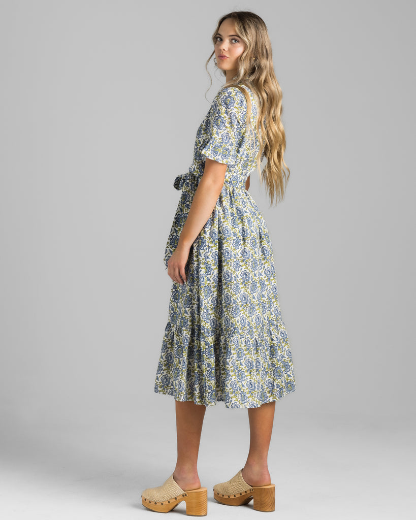 Model wearing a blue and green floral print, short sleeve with ruffles, buttondown, collared, midi length dress.