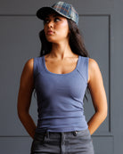 A woman wearing a fitted scoop-neck tank top in dusty periwinkle blue, paired with a plaid flat cap and gray jeans, standing against a dark gray paneled wall.