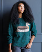 A woman with long, curly black hair wearing a dark teal-green knit sweater with a large duck motif across the chest, paired with dark gray jeans, standing against a dark blue paneled wall.
