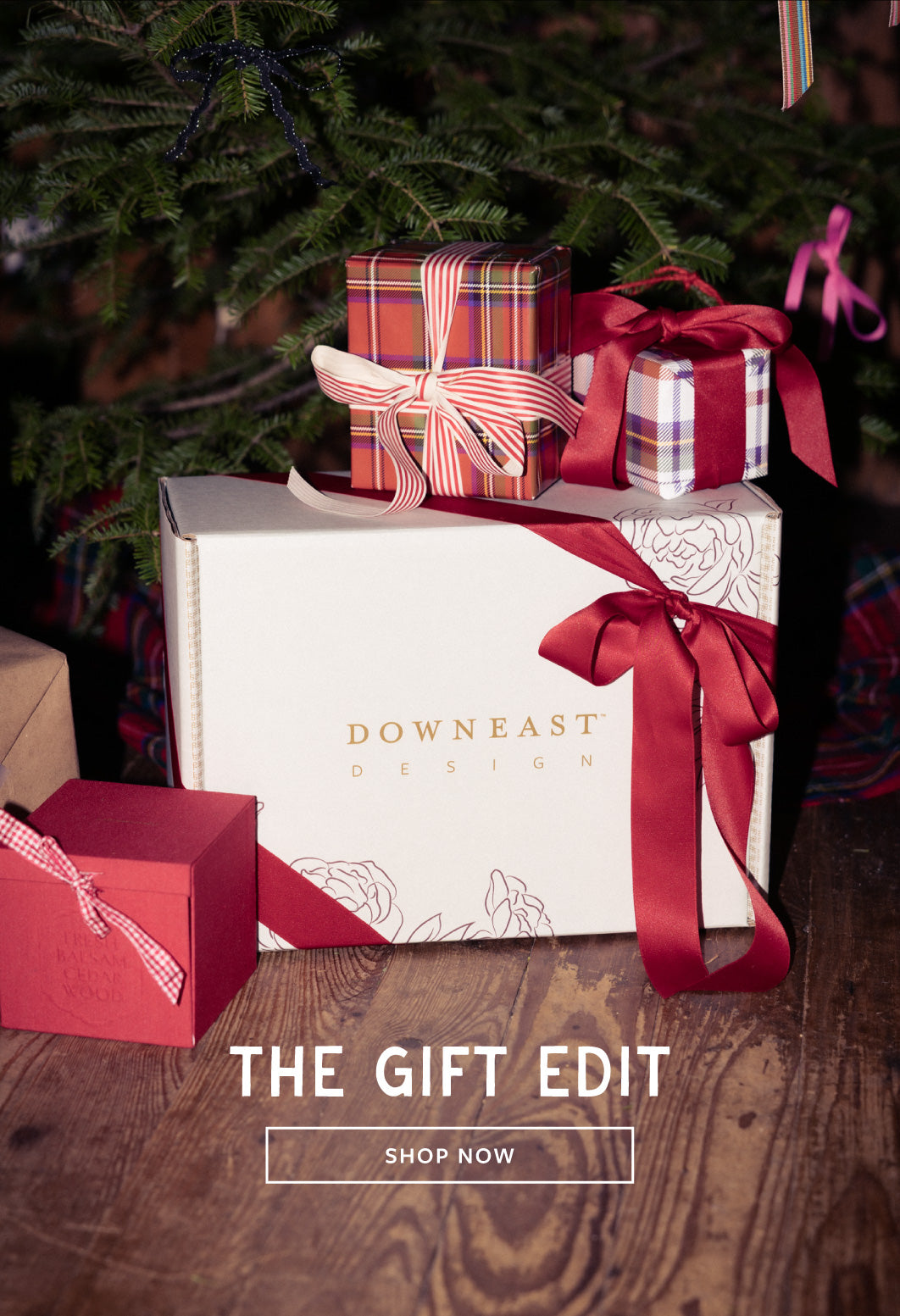 A white Downeast Design gift box with a large red ribbon sits on a wooden surface beside smaller wrapped presents under a Christmas tree. Text at the bottom reads 'THE GIFT EDIT' with a 'SHOP NOW' button.