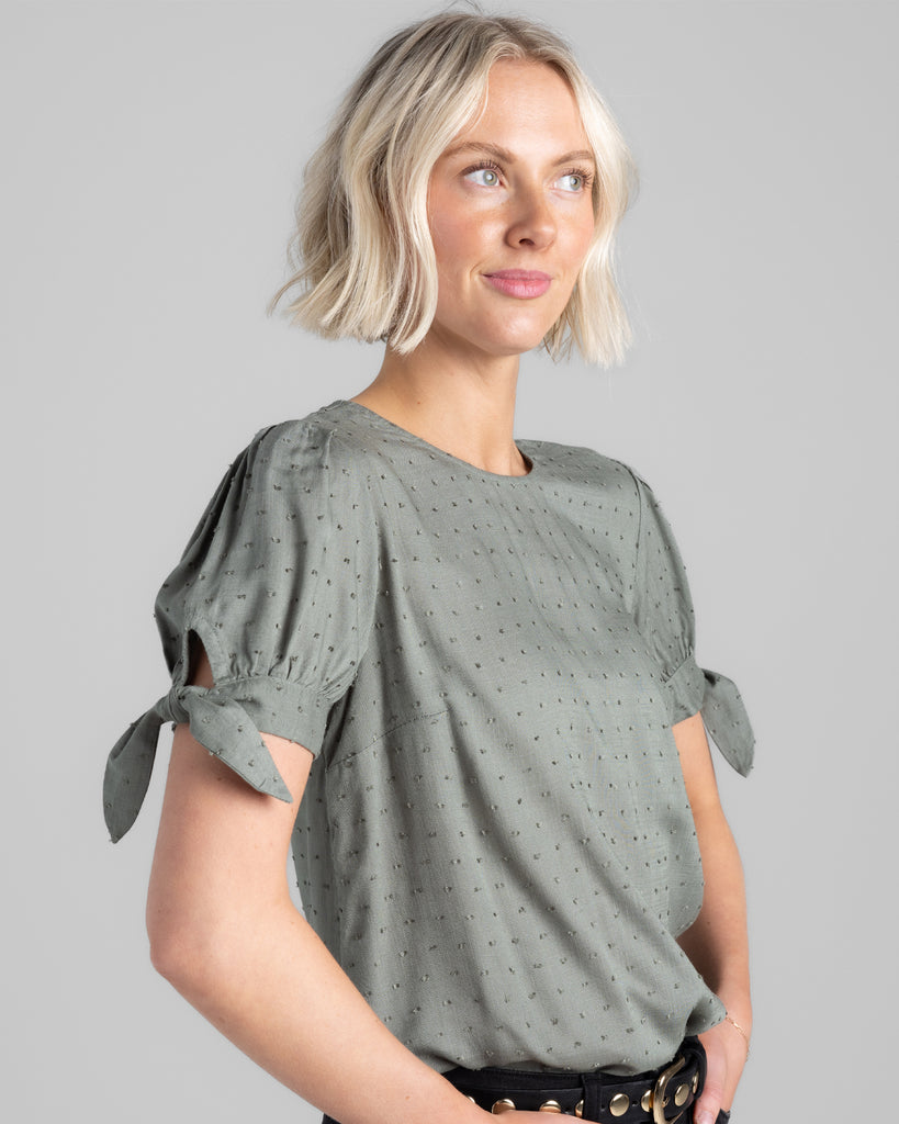 Model wearing a sage green short sleeve with ties, round neck blouse with dotted swiss texturing.