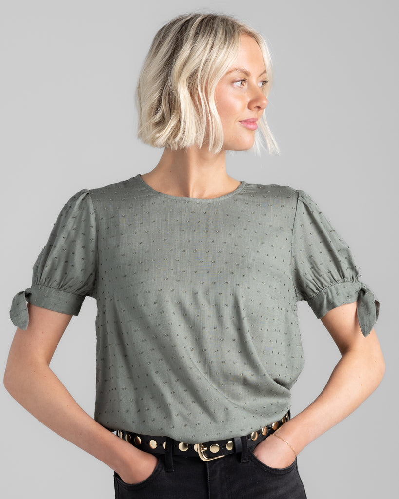 Model wearing a sage green short sleeve with ties, round neck blouse with dotted swiss texturing.