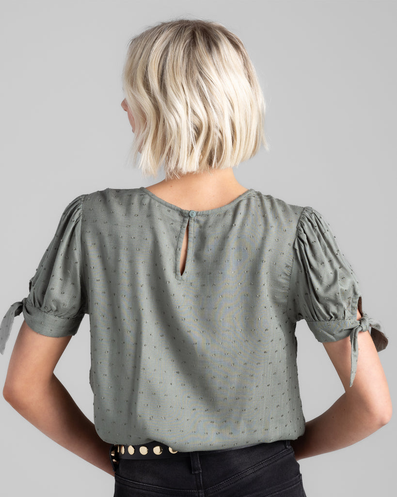 Model wearing a sage green short sleeve with ties, round neck blouse with dotted swiss texturing.