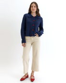 Woman wearing a navy blue cardigan with red buttons, beige pants, and red shoes on a white background