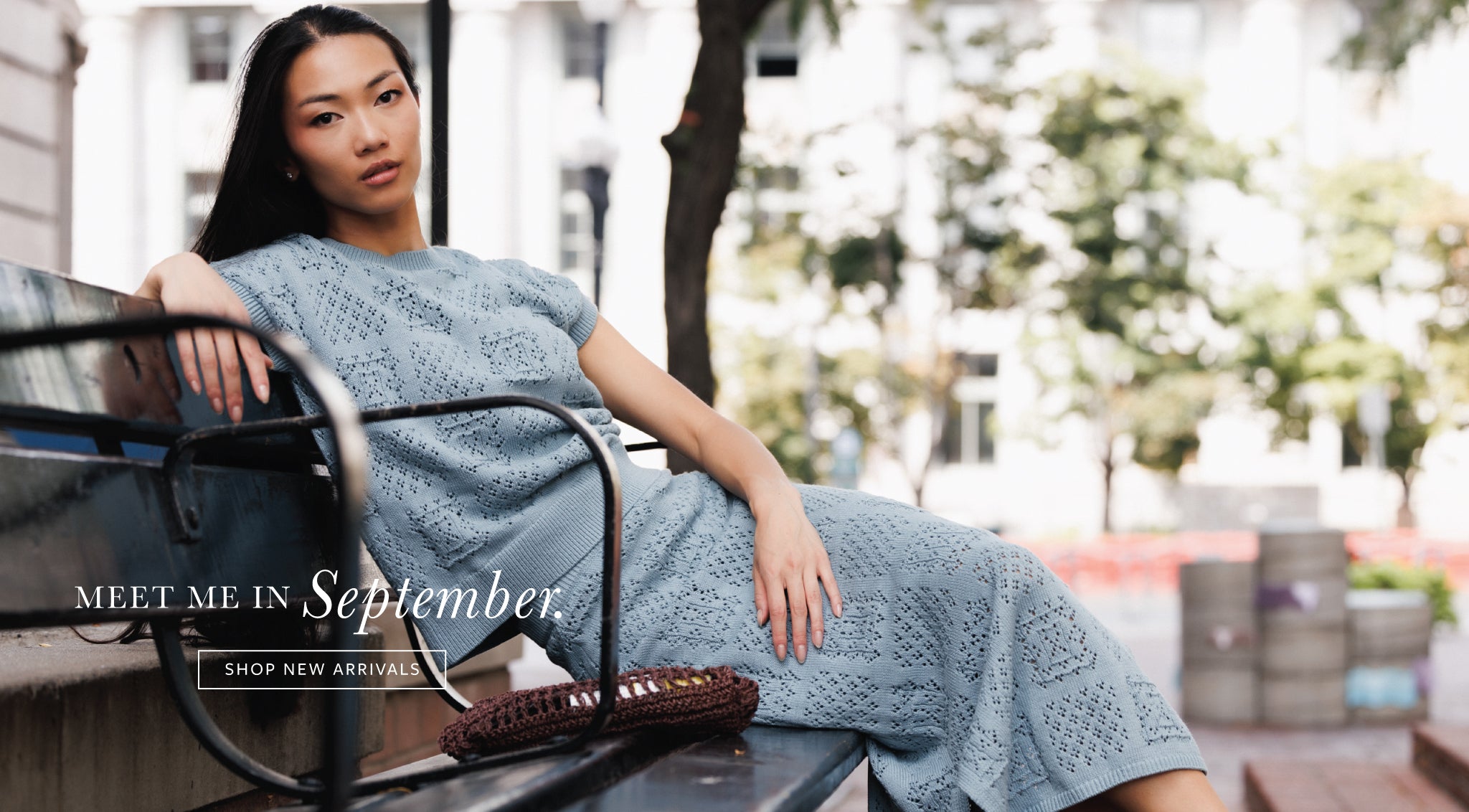A woman reclining on a bench outdoors wearing a light blue textured knit dress, with text overlay reading 'MEET ME IN September. SHOP NEW ARRIVALS'.
