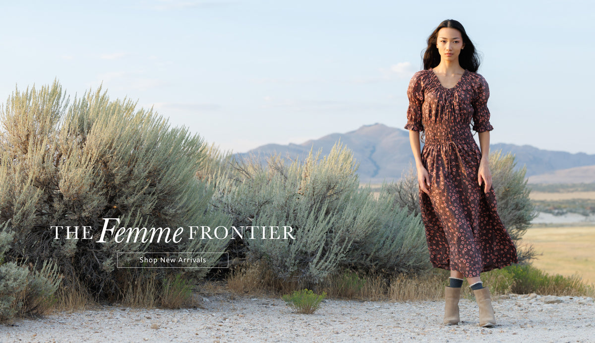 A woman stands outdoors in a desert landscape wearing a brown and black patterned midi dress with puff sleeves and beige ankle boots. Large text on the left reads 'THE Femme FRONTIER' with a smaller 'Shop New Arrivals' line.
