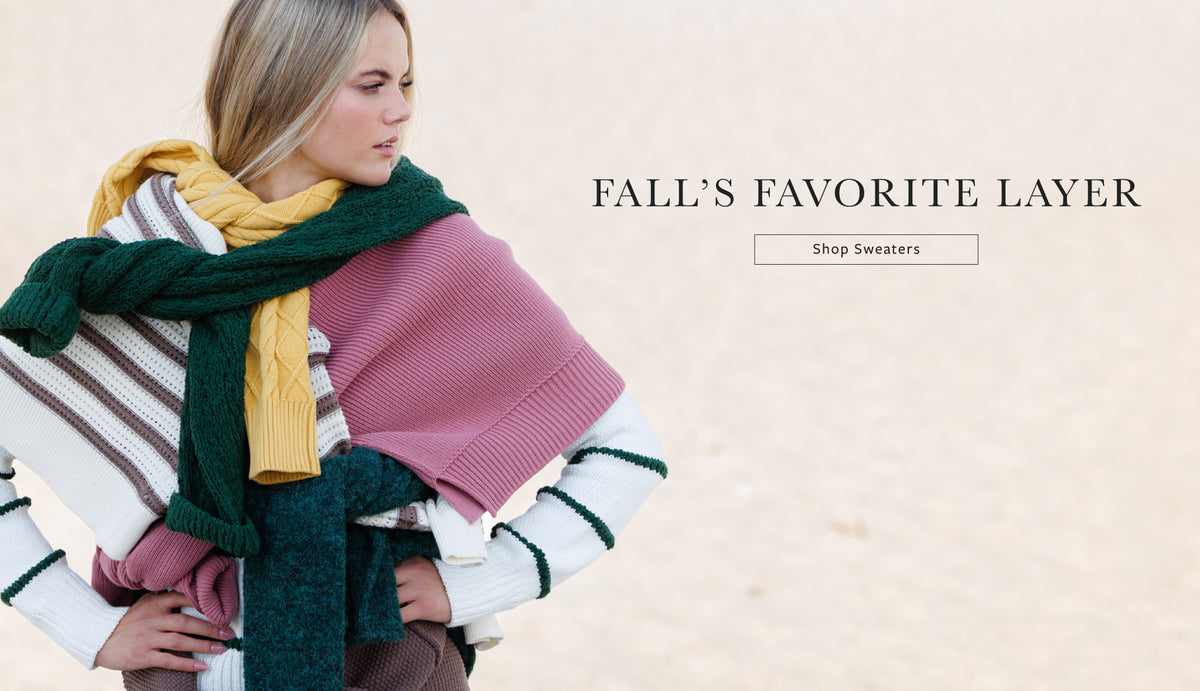 A woman wearing a pink knit sweater layered with a striped white sweater and wrapped in a dark green and yellow scarf, set against a light beige background with the text 'FALL'S FAVORITE LAYER' and a 'Shop Sweaters' button.