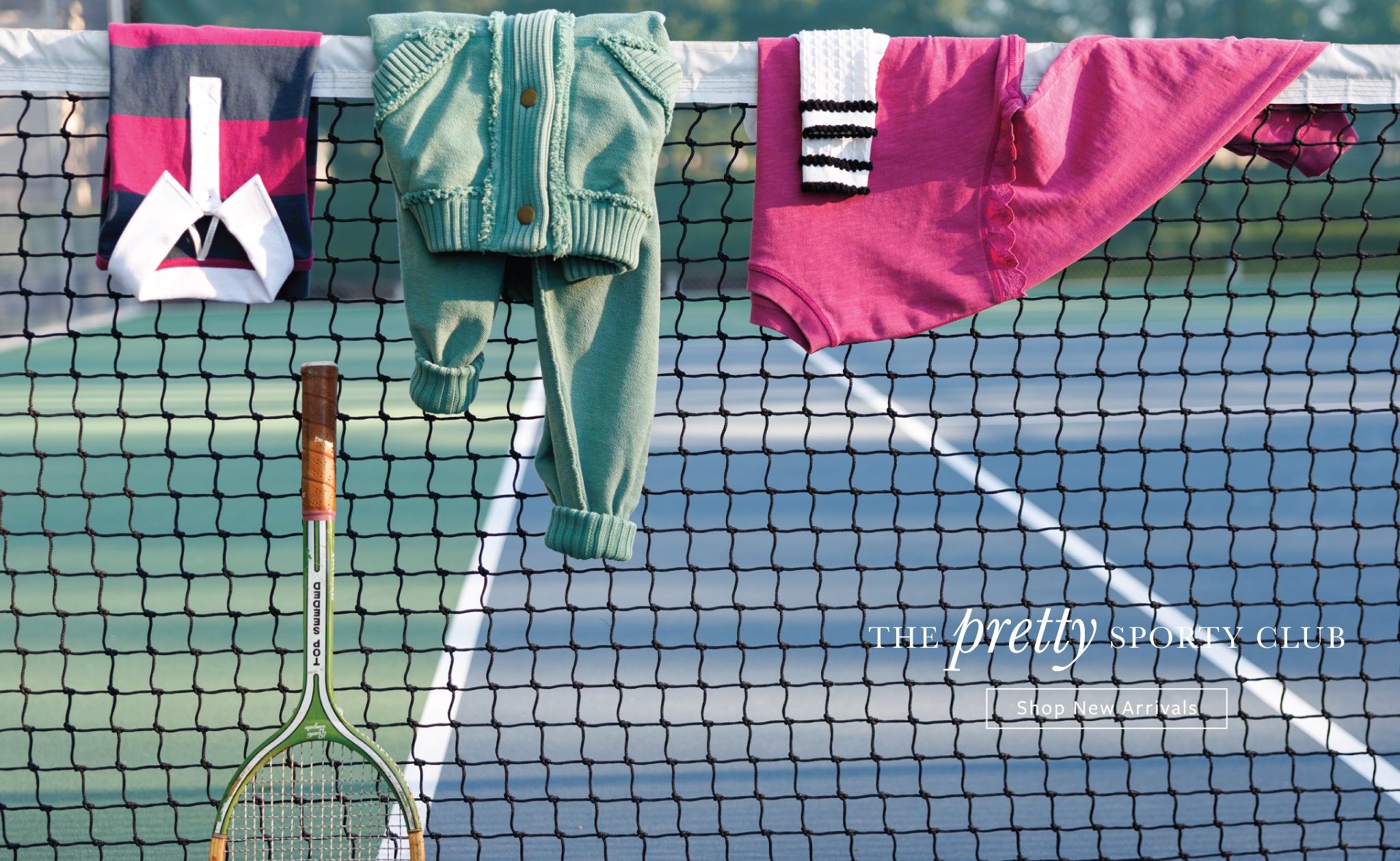 A lifestyle shot of a tennis court net with sports apparel hanging on it. A mint green knit cardigan and matching pants are hanging in the center, flanked by a bright pink sweatshirt and a dark pink and white towel.