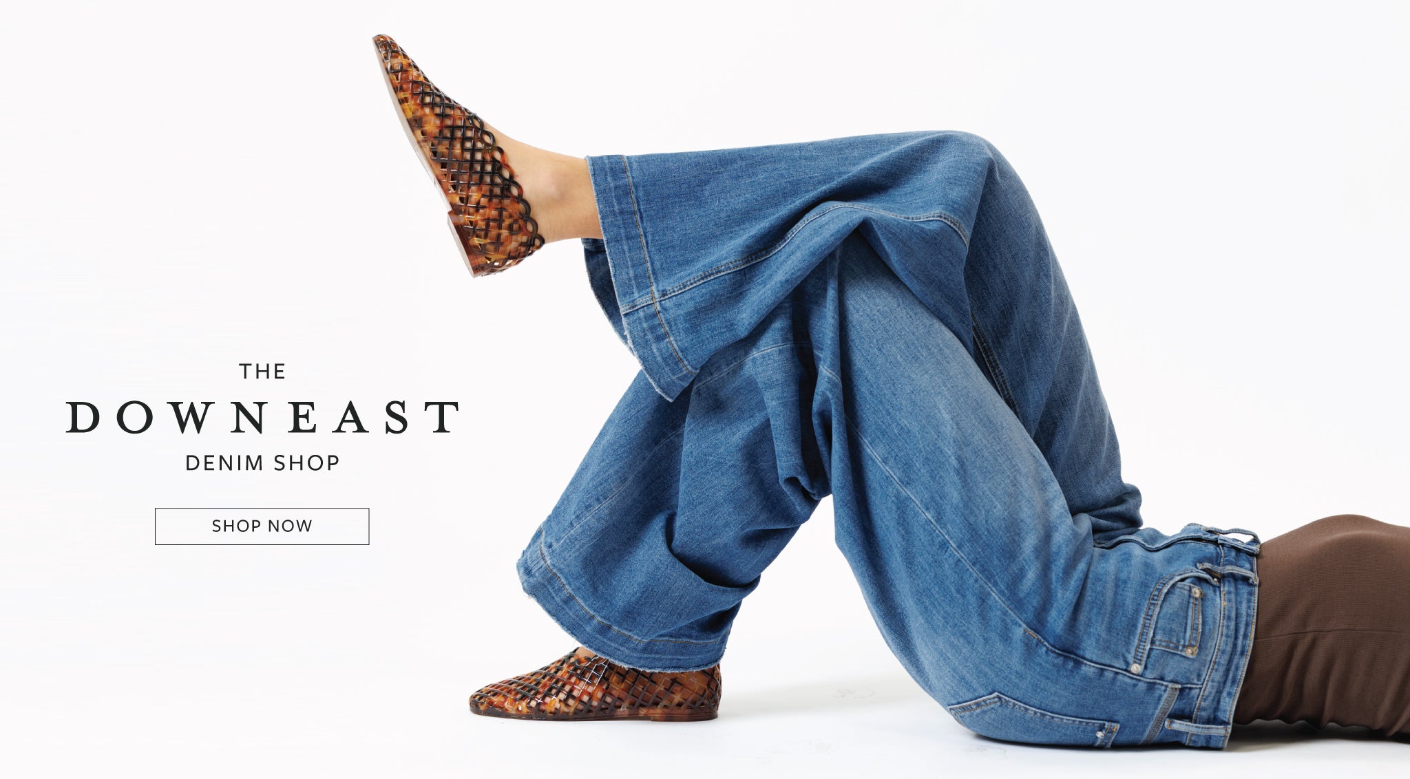 A person reclining on a white studio floor wearing loose blue jeans and leopard-print slip-on shoes, with a visible 'THE DOWNNEAST DENIM SHOP SHOP NOW' text overlay on the left.