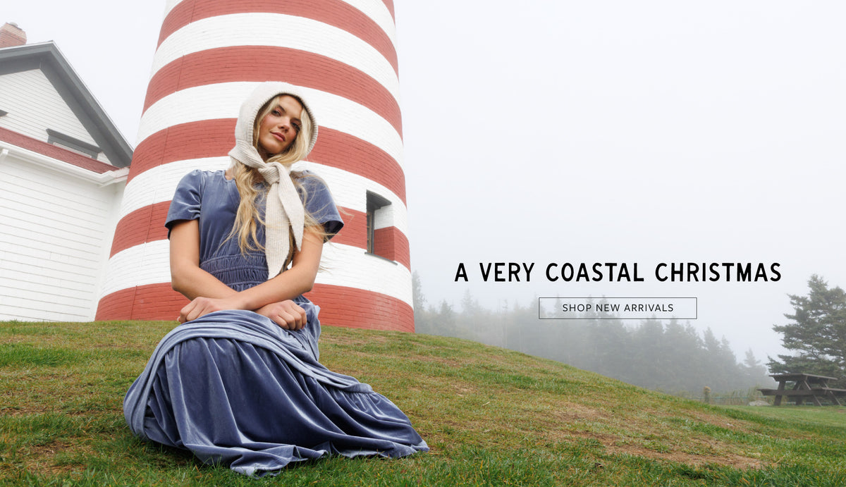 A woman in a vintage-style blue dress and white bonnet sits on a grassy hill in front of a red-and-white striped lighthouse, with misty trees in the background. Text on the right reads 'A VERY COASTAL CHRISTMAS' and 'SHOP NEW ARRIVALS'.