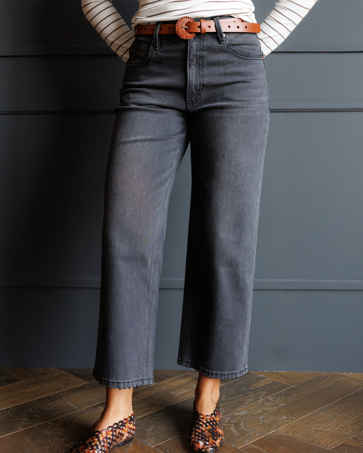 A person wearing dark gray wide-leg cropped jeans with a brown belt, a cream striped long-sleeve top, and patterned flat shoes, standing against a dark gray paneled wall on a wood floor.
