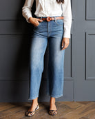 A model wearing high-waisted, wide-leg cropped blue jeans with a frayed hem, styled with a white blouse, a brown leather belt, and leopard-print flat shoes, standing against a dark gray paneled wall.