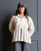 A woman wearing a cream-colored lace-trimmed zip-up hoodie with a peplum hem, paired with gray pants and a plaid cap, standing against a dark gray paneled wall.