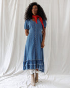 A woman stands against a white draped fabric backdrop wearing a short-sleeve blue denim midi dress with a button front, a red neck scarf, white tights, and beige ankle boots.