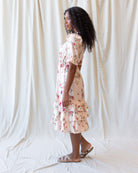 A woman wearing a short-sleeve cream-colored midi dress with a pink and red floral print, standing in profile against a draped white fabric backdrop.