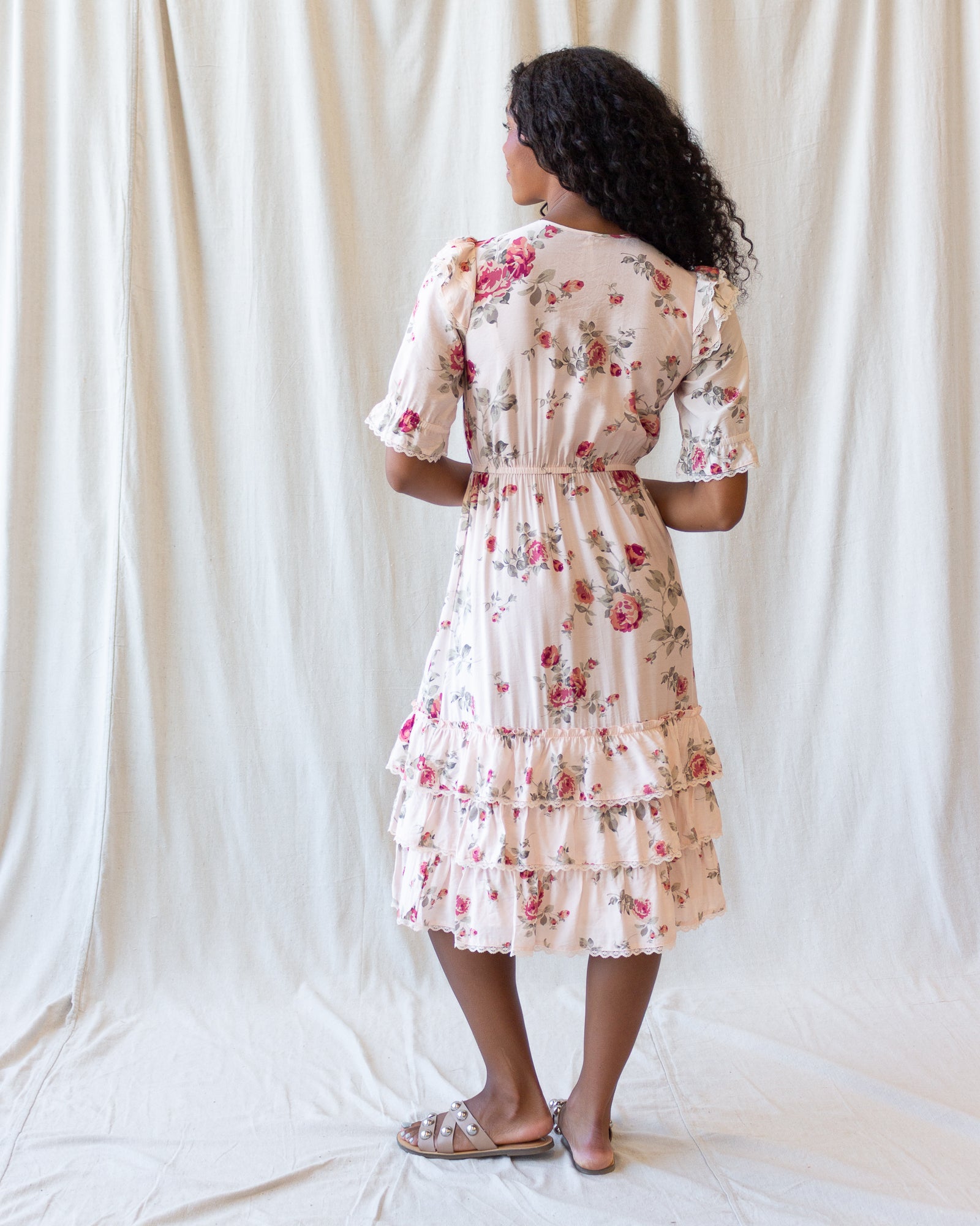 A woman stands with her back to the camera wearing a short-sleeve tiered midi dress with a pink and green floral print on a cream background, paired with silver sandals, set against a draped white fabric backdrop.