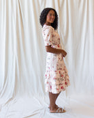 A woman wearing a short-sleeve tiered midi dress with a pink and red floral print on a cream background, standing in front of a white fabric backdrop.
