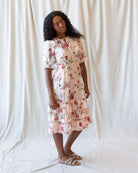 A woman modeling a cream-colored tiered midi dress with a red and green floral print, standing against a draped white fabric backdrop.