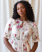 A woman with curly black hair wearing a cream-colored short-sleeve dress with a pink and green floral print, lace ruffle details, and a tie-neck design, posed against a soft beige curtain background.