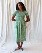 A woman modeling a light green midi dress with a white floral pattern, standing against a draped white fabric backdrop.