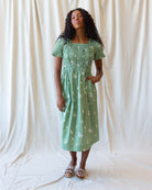 A woman modeling a sage green midi dress with white floral embroidery, standing against a draped white fabric backdrop.