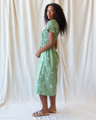 A woman stands in profile wearing a light mint green midi dress with a white floral pattern, short puff sleeves, and a gathered waist. She is posed against a draped cream fabric backdrop.