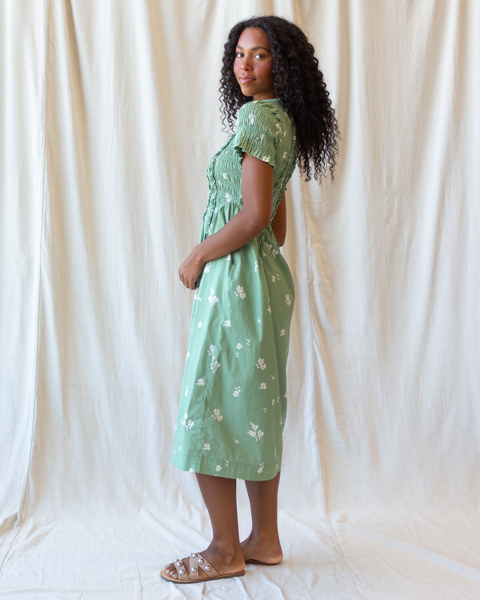 A woman stands in profile wearing a light mint green midi dress with a white floral pattern, short puff sleeves, and a gathered waist. She is posed against a draped cream fabric backdrop.