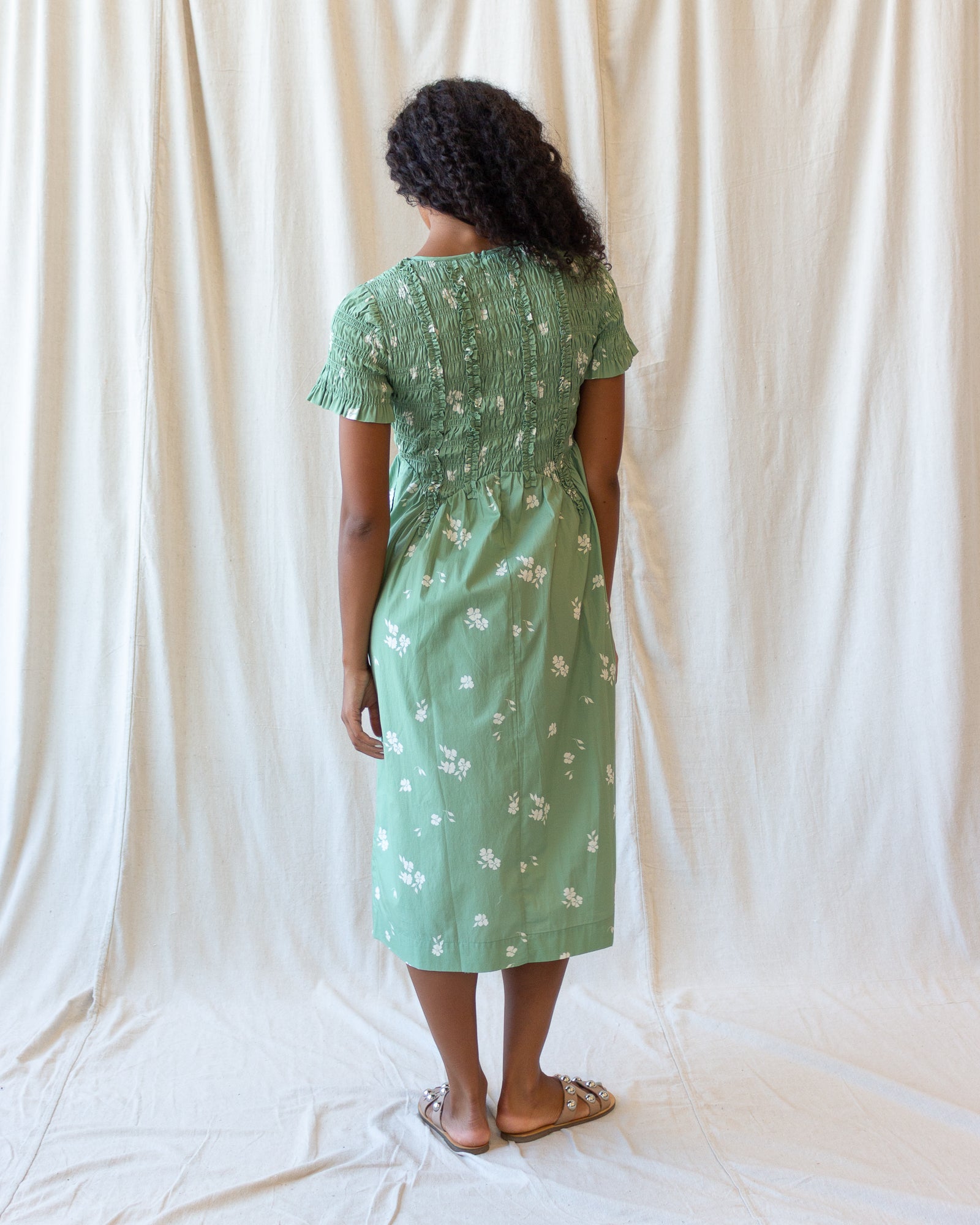 A woman stands with her back to the camera wearing a sage green midi dress with white floral embroidery and ruffled short sleeves, set against a draped white fabric backdrop.