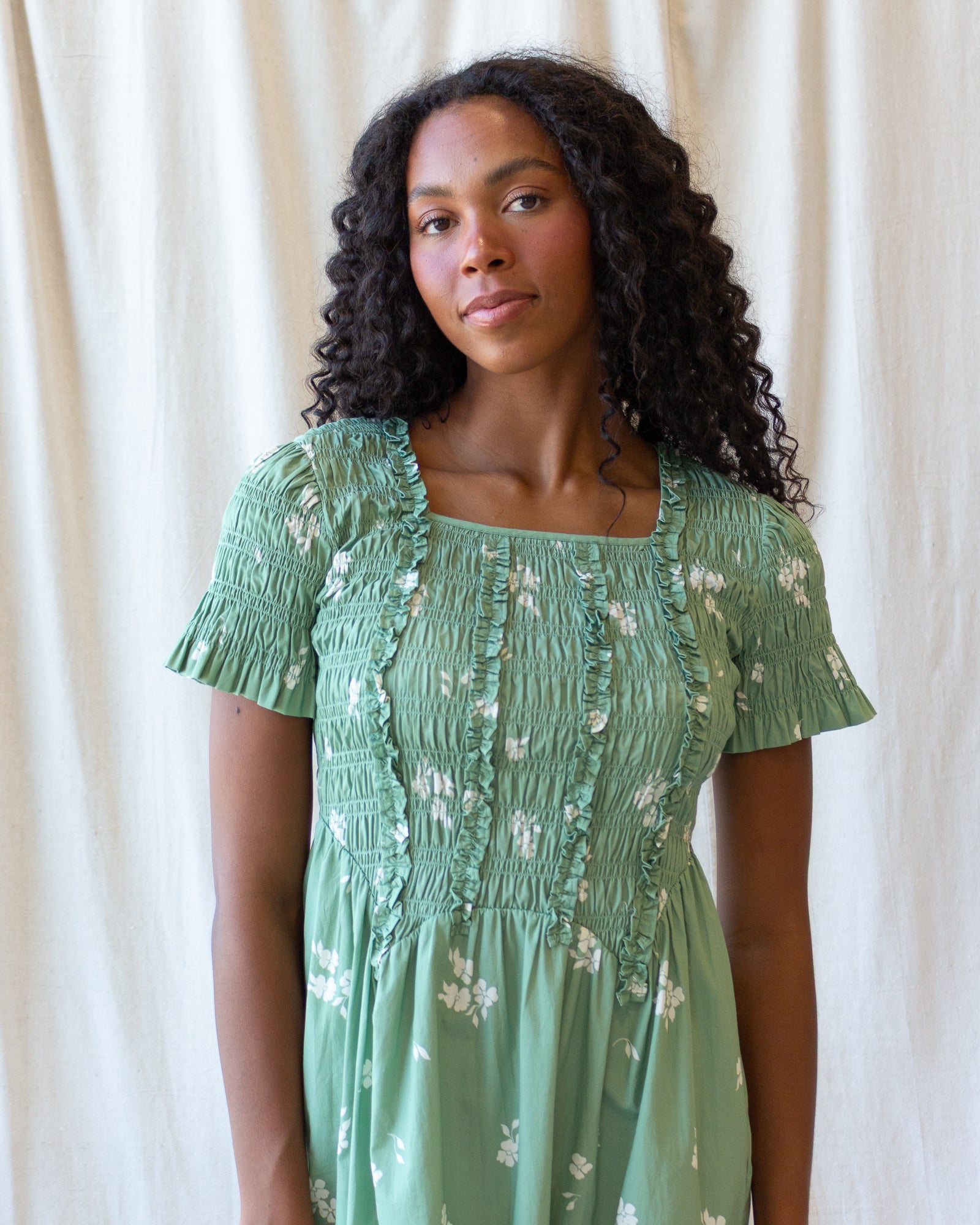 A woman with long, curly black hair wearing a sage green smocked dress with white floral patterns, standing against a draped cream fabric backdrop.