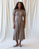 A woman stands against a draped off-white fabric backdrop wearing a short-sleeve, button-front midi dress in a muted taupe-brown color. The dress has a collared neckline and a textured, slightly wavy fabric.