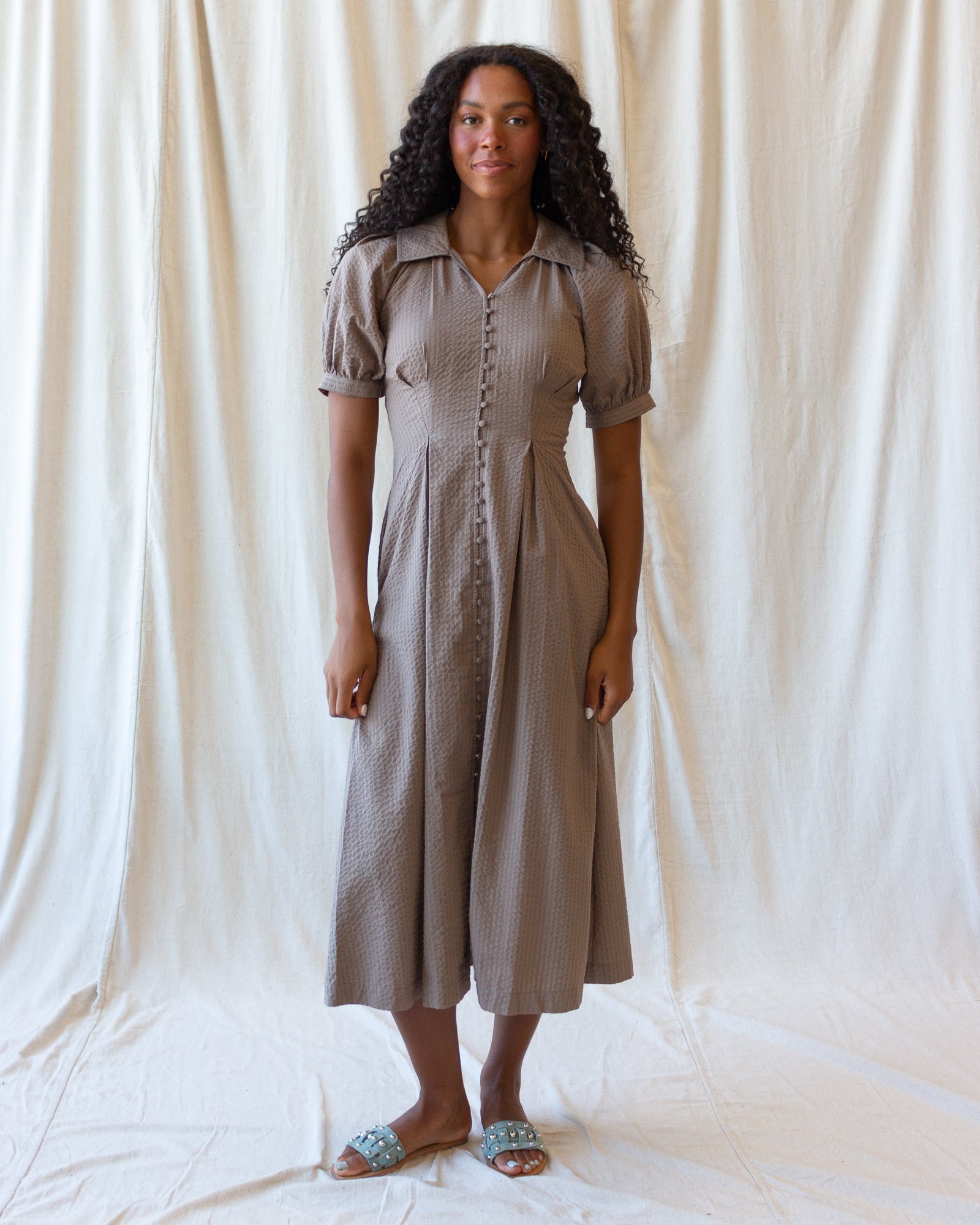 A woman stands against a draped off-white fabric backdrop wearing a short-sleeve, button-front midi dress in a muted taupe-brown color. The dress has a collared neckline and a textured, slightly wavy fabric.