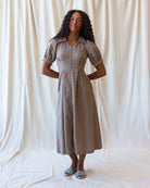A woman modeling a taupe-brown short-sleeve midi dress with puff sleeves, a button-down front, and a gathered waist, standing against a draped white fabric backdrop.