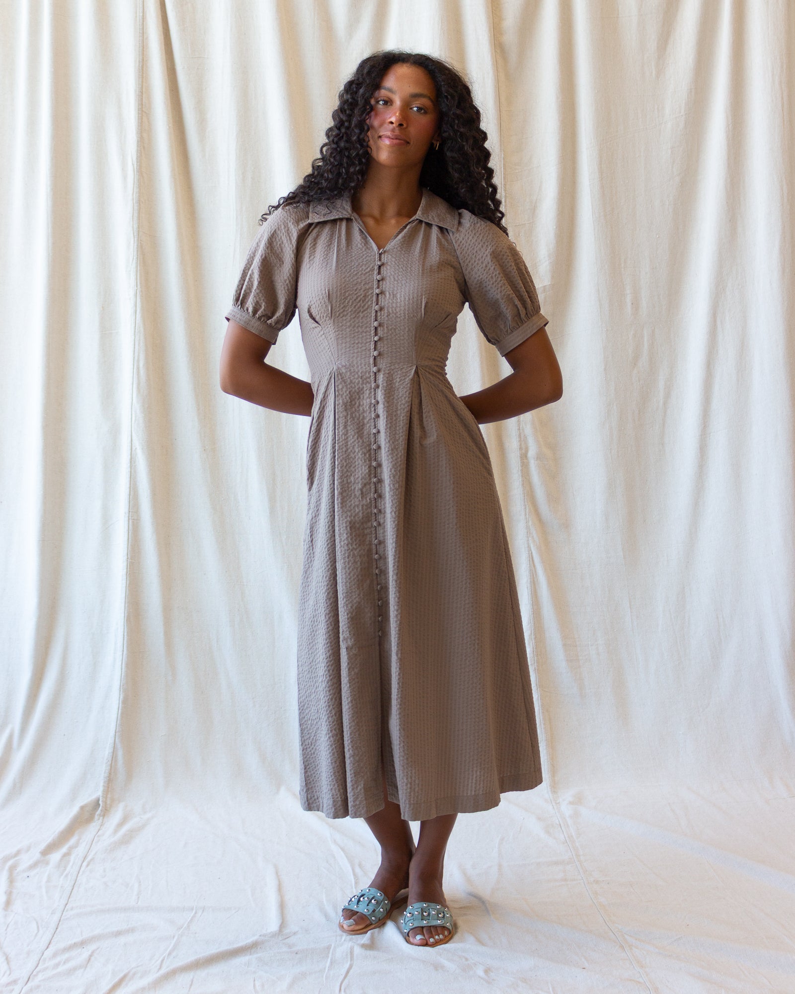 A woman modeling a taupe-brown short-sleeve midi dress with puff sleeves, a button-down front, and a gathered waist, standing against a draped white fabric backdrop.