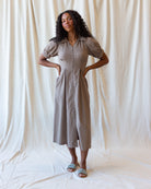 A woman modeling a taupe-colored midi dress with puff sleeves, a button-down front, and side pockets, standing against a draped cream fabric backdrop.